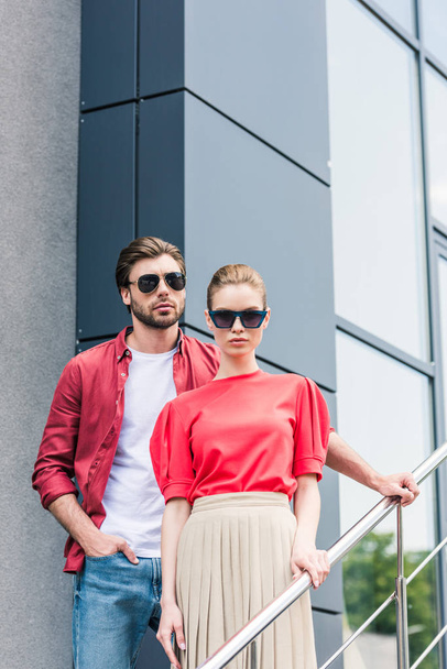 front view of stylish young couple of models in sunglasses standing on staircases  - 写真・画像