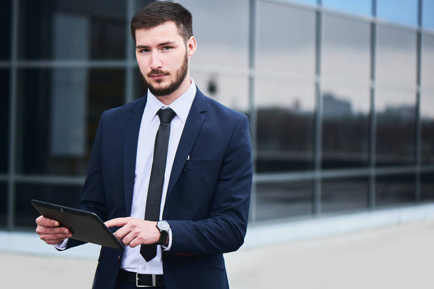 handsome businessman looking at camera while using tablet pc with glass building on background  - Foto, Bild
