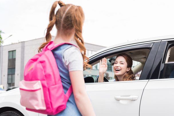 enfoque selectivo de la mujer sonriente en el coche saludando a la hija con la mochila
 - Foto, Imagen