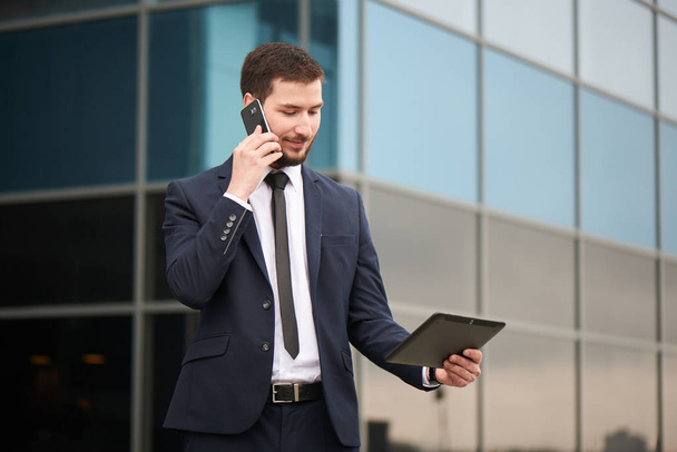 businessman talking on phone and looking at tablet pc in hand with glass building on background  - Foto, imagen