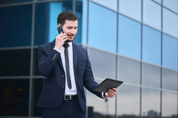 businessman talking on phone and looking at tablet pc in hand with glass building on background  - Foto, Imagen