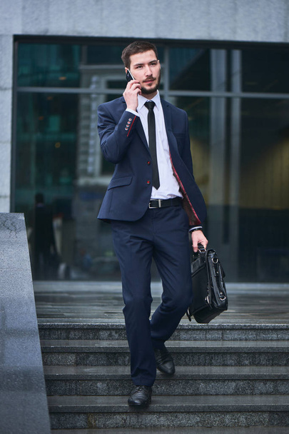 serious handsome businessman in suit and white shirt with briefcase in hand talking on phone while descending steps  - Foto, Imagen