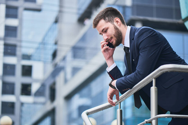 handsome businessman in suit  talking on phone while leaning on railing with building on background - Φωτογραφία, εικόνα
