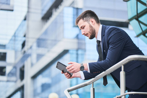 serious elegant businessman using tablet pc while standing on porch of business center - Foto, imagen