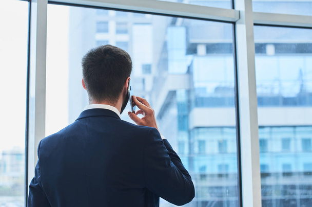 bearded businessman talking on phone and looking at cityscape outside window - Фото, изображение