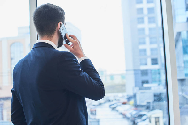 bearded businessman talking on phone and looking at cityscape outside window - Photo, image