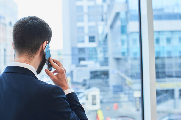 bearded businessman talking on phone and looking at cityscape outside window - Фото, зображення