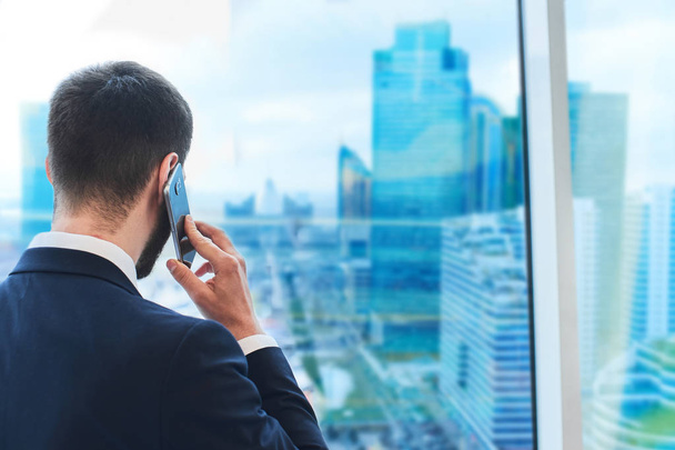 bearded businessman talking on phone and looking at cityscape outside window - Foto, immagini