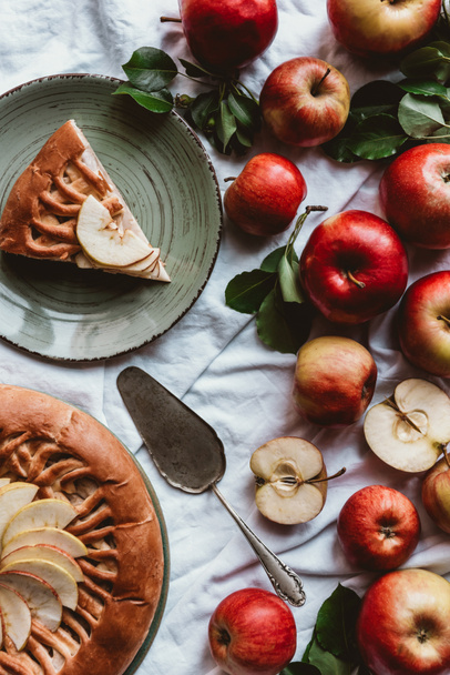 flache Lage mit Apfelkuchen, Tortenserver, frischen Äpfeln und grünen Blättern auf weißer Tischdecke - Foto, Bild