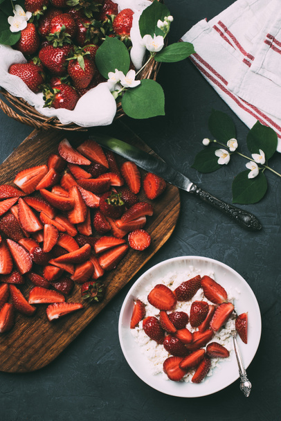 top view of sliced strawberries on wooden cutting board, cottage cheese and jasmine flowers on black  - Foto, Bild
