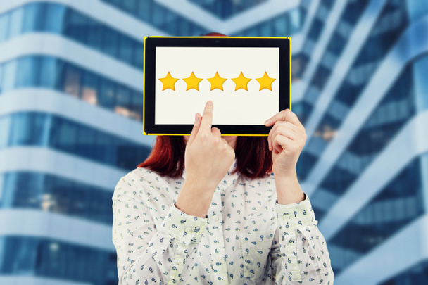 Young businesswoman covering her face using a digital tablet with five golden stars on the screen trying to choose a feedback. Excellent customer service rating concept. - Photo, Image
