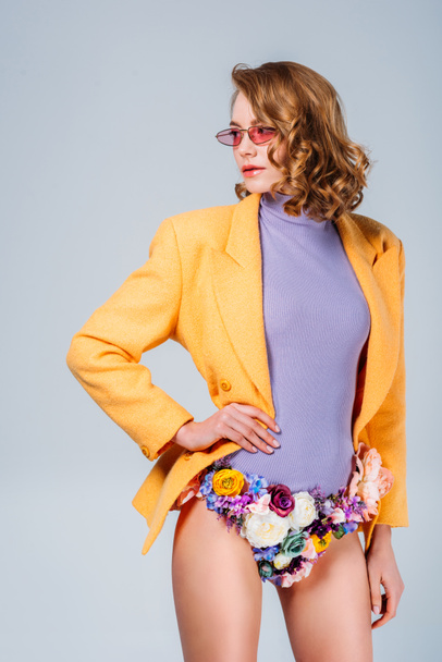 girl in sunglasses and panties made of flowers standing with hand on waist and looking away isolated on grey - Photo, Image