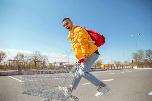 fashion guy in sunglasses and a yellow jacket jump in a cart from food in the supermarket parking - Fotografie, Obrázek