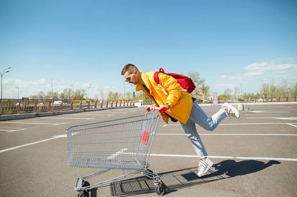 fashion guy in sunglasses and a yellow jacket a cart from food in the supermarket parking - Photo, image