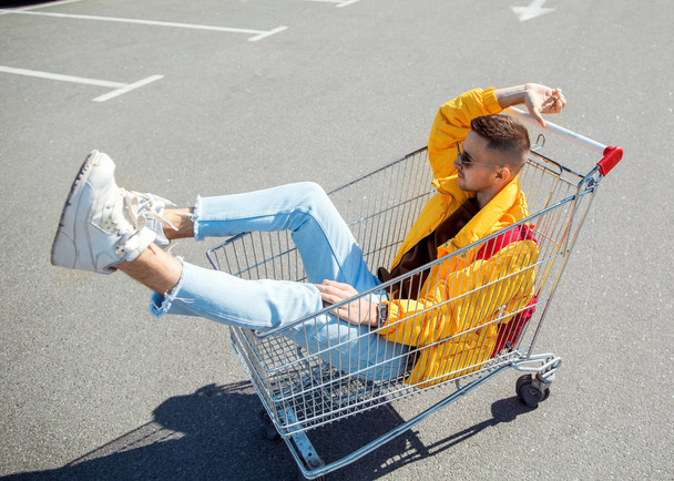 fashion guy in sunglasses and a yellow jacket sits in a cart from food in the supermarket parking - Фото, изображение