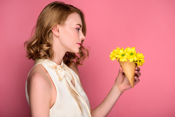 side view of beautiful girl holding waffle cone with yellow flowers on pink - Фото, зображення