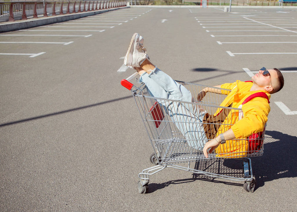 fashion guy in sunglasses and a yellow jacket sits in a cart from food in the supermarket parking - Photo, Image