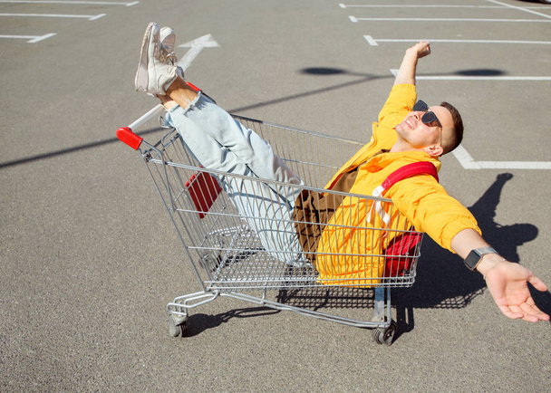 fashion guy in sunglasses and a yellow jacket sits in a cart from food in the supermarket parking - 写真・画像