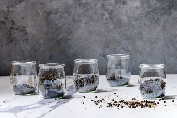 Variety of different black peppers allspice, pimento, long pepper, monks pepper, peppercorns and ground powder in glass jars on white marble kitchen table. - Φωτογραφία, εικόνα