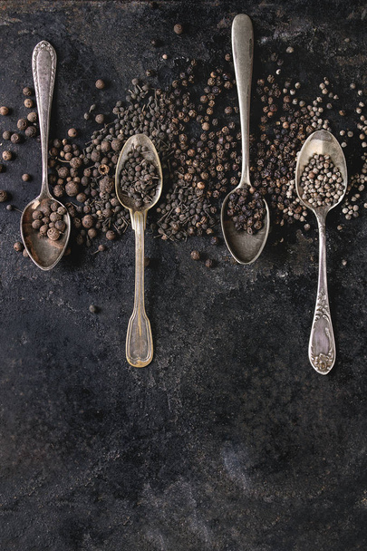 Variety of different black peppers allspice, pimento, monks pepper, peppercorns in vintage spoons over old black iron texture background. Top view, space. - Φωτογραφία, εικόνα