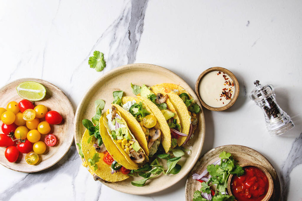 Variety of vegetarian corn tacos with vegetables, green salad, chili pepper served on ceramic plate with tomato and cream sauces with ingredients above over white marble background. Top view, space. - Foto, Imagem