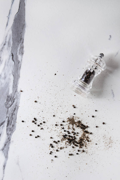 Black peppers peppercorns and ground powder from transparent pepper mill over white marble texture background. Top view, space. - Photo, Image