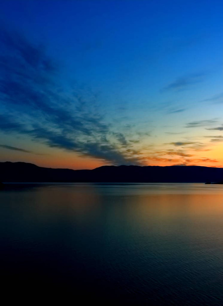 Narancsszín napkelte tükrözi a sötét nyugodt vízfelület a Bajkál-tó kora reggel. Nyári táj napkelte felett silhouette hegylánc a tó partján egy másik vízzel - Fotó, kép