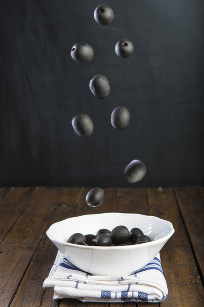 Black olives falling in a bowl on a wooden table - Foto, imagen