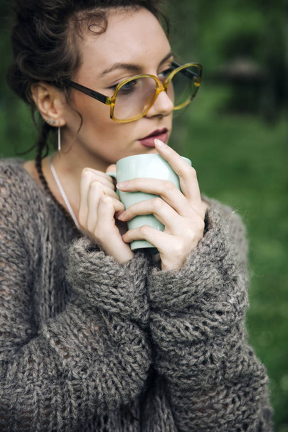 Portrait de jeune femme en pull et avec des lunettes buvant du café dans le parc
 - Photo, image