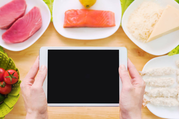Design workpiece. Female hands hold a tablet PC over a table with raw seafood cheese and vegetables. Blank display for presentation on the topic of food - Fotó, kép