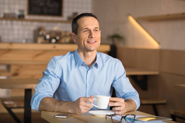 Thoughtful smiling businessman taking break and drinking coffee in cafe - Photo, image