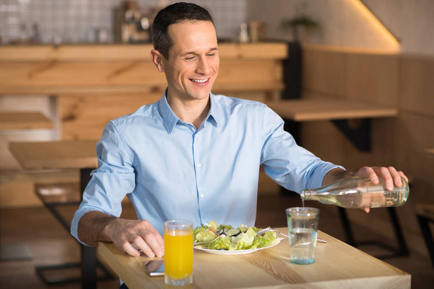 Lächelnder Geschäftsmann gießt beim Mittagessen im Café Wasser aus der Flasche ins Glas - Foto, Bild