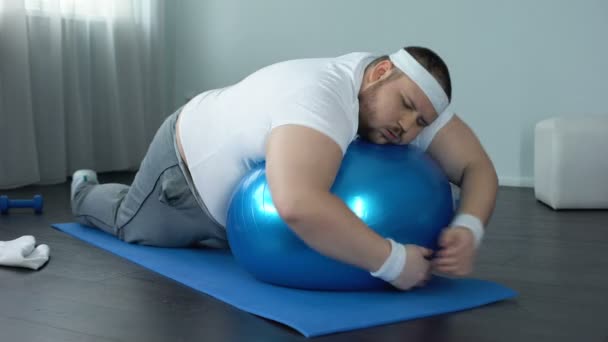 Weak-willed fat man relaxing on fitness ball, home workout break, laziness - Záběry, video