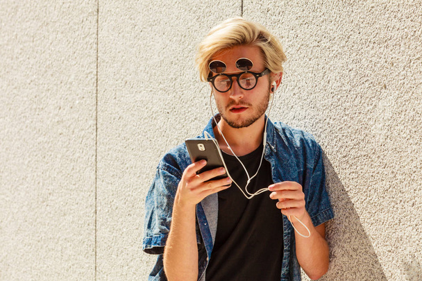 Men fashion, technology, urban style clothing concept. Hipster guy standing on city street wearing jeans outfit and eccentric sunglasses listening to music and holding phone - Photo, Image