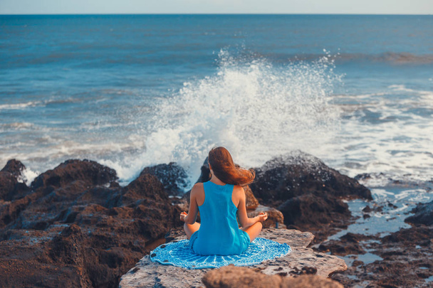 Frau praktiziert Yoga auf Klippe bei stürmischem Ozean. Rückansicht. blauer Himmel. Konzept eines gesunden Lebensstils. - Foto, Bild