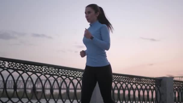 sporty girl in a smart watch jogs along the embankment at sunset. portrait of a sportswoman taking a sport in nature - Video