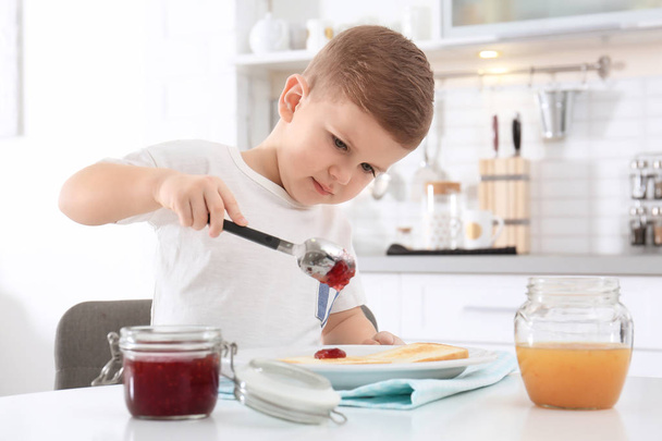 Cute little boy spreading sweet jam onto toast at table - Photo, Image