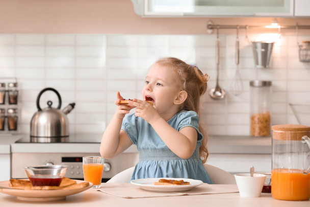 Adorable little girl eating tasty toasted bread with jam at table in kitchen - Foto, afbeelding