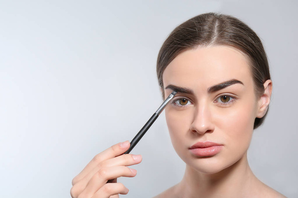 Young woman correcting shape of eyebrow with brush on light background - 写真・画像