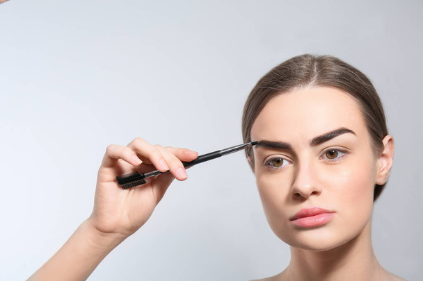 Young woman correcting shape of eyebrow with brush on light background - Photo, image