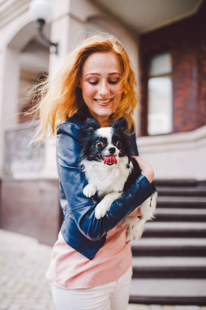 A beautiful young woman with red long hair is holding a small, cute funny big-eyed dog of two flowers, a black-and-white pet of the breed of hichuahua against a house of red brick in summer. - Foto, afbeelding