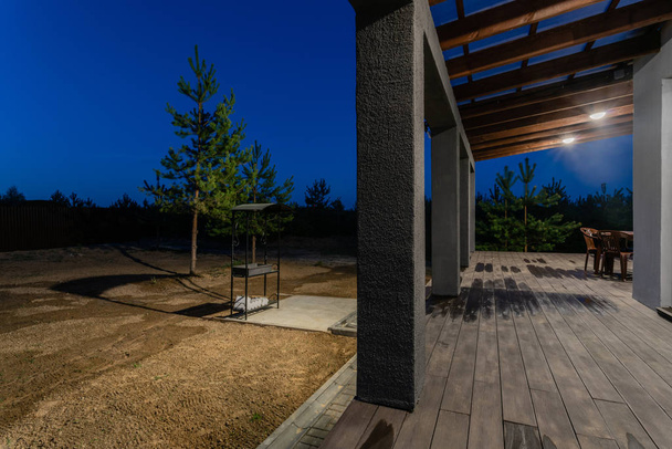 Vista lateral de una veranda abierta frente a una casa de campo forestal moderna. Bosque joven de pino bajo los rayos del atardecer en el fondo
. - Foto, imagen
