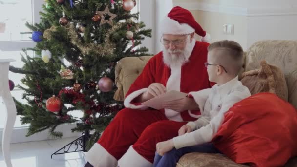 Little boy in stripped jacket and Santa Claus are sitting at the sofa in living room. Child gives envelop to graybeard. Old man opens envelope and reads Christmas letter with emotions of surprise. - Video, Çekim