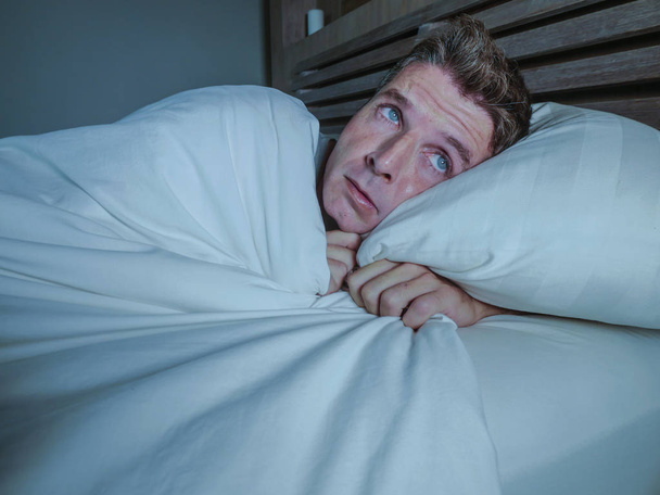 portrait of young attractive scared man in fear and panic suffering horror nightmare covering face with blanket sleepless at night lying on bed in dark bedroom in sleeping disorder concept - Photo, image