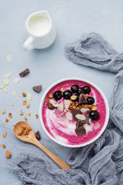 Pink black currant berries, banana smoothie bowl with granola, flakes from almonds and chocolate on light gray concrete background. Selective focus. Top view. Copy space. - Foto, imagen