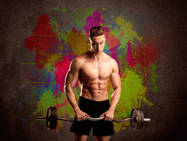 A muscular young bodybuilder lifting weight and showing his hot body with muscles in front of an urban painted wall concept - Foto, imagen