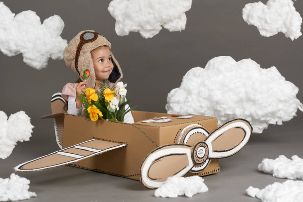 a menina brinca em um avião feito de caixa de papelão e sonhos de se tornar um piloto, nuvens de lã de algodão em um fundo cinza
 - Foto, Imagem