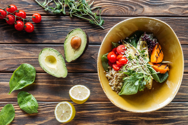 flache Lage mit vegetarischem Salat mit gegrilltem Gemüse, Rosenkohl, Kirschtomaten in Schüssel und frischen Zutaten herum auf hölzerner Tischplatte angeordnet - Foto, Bild
