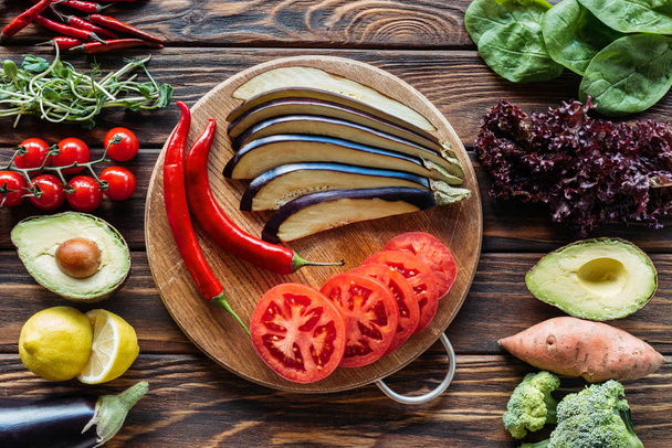 flat lay with cut eggplant, tomatoes and chili peppers on cutting board with fresh vegetables around on wooden surface - 写真・画像