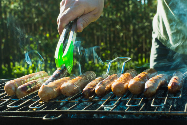 Male hand turned the sausages over an open fire barbecue. Close up. Outdoor. - Photo, Image